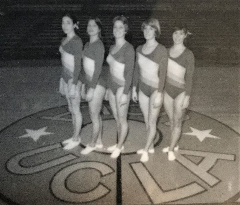 UCLA womens gymnastics teammates standing on the UCLA logo at center court