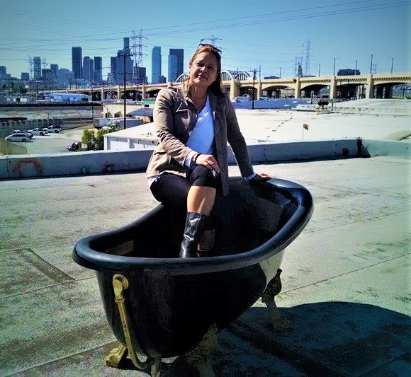 Cheryl Quintana Leader seated in a clawfoot bathtub on a rooftop with the downtown Los Angeles skyline and Sixth Street Bridge behind her